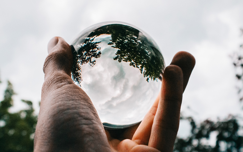 Main tenant une sphère en verre réfléchissante montrant une vue inversée des arbres et d'un ciel nuageux.